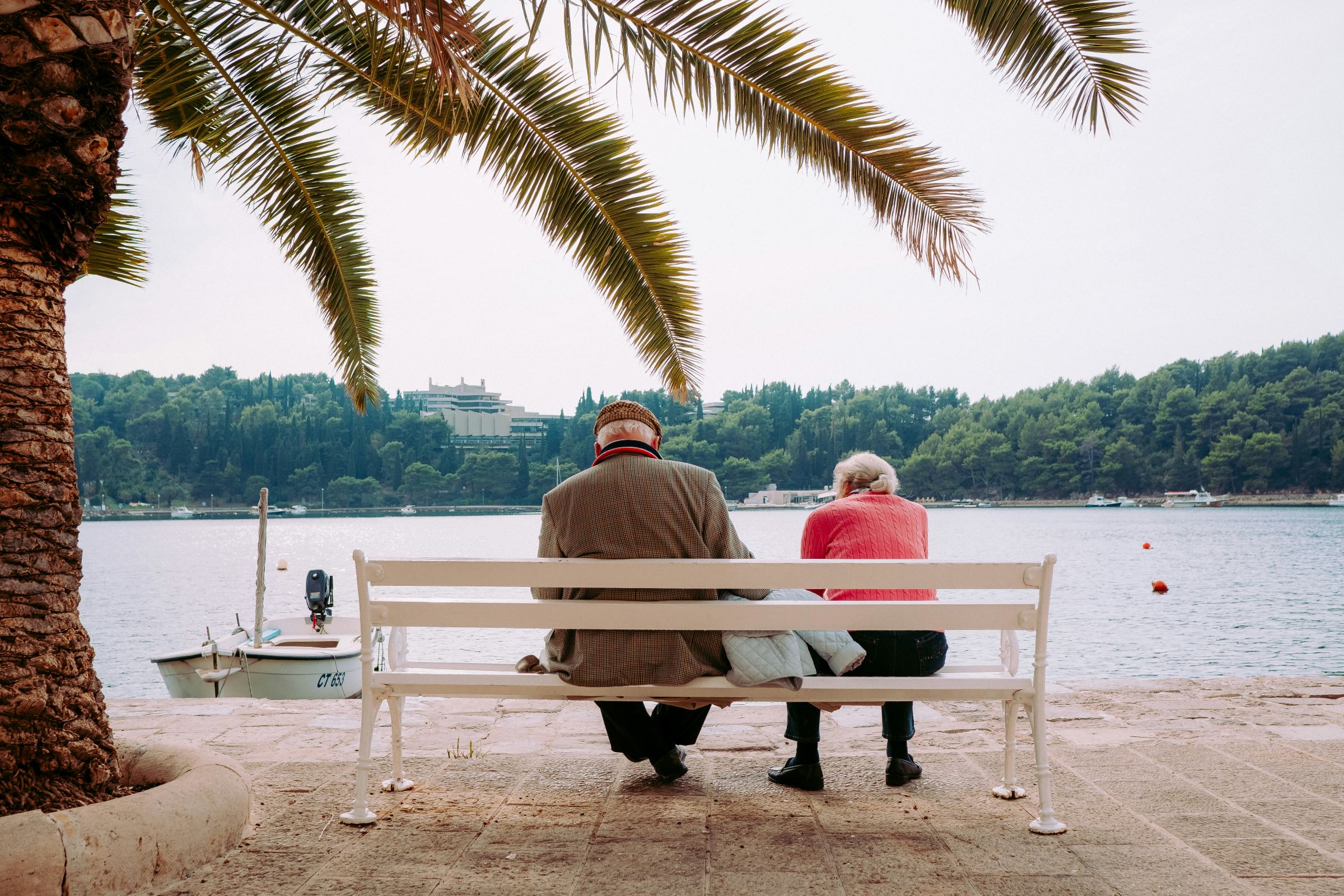 Retired couple sit on a bench by a palm tree, overlooking a lake with boats.